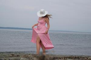 vit hatt flicka i en rosa klänning på de hav en bred skott av en tonåring promenader barfota på de strand foto