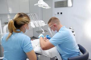 få tänder rengöring och putsning på klinik tandläkare arbetssätt på patient i stol. läkare i blå handskar gäller salva till patientens tänder den där visar var där är plack den där behov till vara rengöras. foto