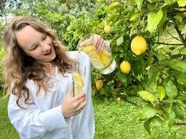 dess bara en citron- paradis, en Tonårs flicka står mot de bakgrund av en citron- träd med citroner och drycker citronsaft från en glas glas i henne Övrig hand, hon innehar en kanna av dryck foto