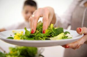 matlagning sallad mamma med hand lägger till sallad löv till stor skål i bakgrund pojke rör om sallad kök matlagning förkläden matlagning hemlagad mat kommunikation med barn portion barn i matlagning Träning foto