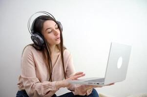 attraktiv Lycklig ung flicka studerande studerar på de högskola bibliotek, Sammanträde på de skrivbord, använder sig av bärbar dator dator, har chatt, vinka foto