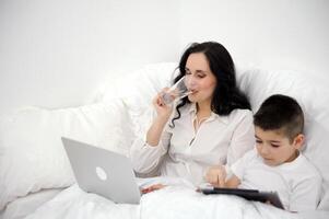 mamma och son i vit säng med läsplatta och bärbar dator mamma dricka i en glas av ren vatten dämpa hosta bantning dricka rena dryck hälsa tar vård av själv foto