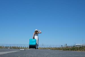 kvinna flicka i vit klänning turist med stor blå resväska går in i distans justeras hatt mot himmel resa bana plats för resa byrå reklam text oigenkännlig människor utvecklande kläder foto