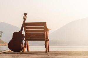 de trä- gitarr var uppsättning upp bredvid en stol på en trä- balkong i de morgon- över en reservoar med skön visningar av natur och ljus solsken i förberedelse för de kommande fest. foto