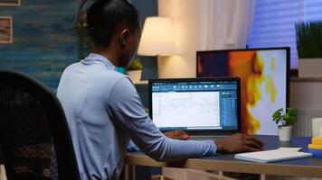 svart anställd kontroll digital blå grafik analyserar projekt av företag ser på bärbar dator Sammanträde på skrivbord i levande rum kontor sent på natt över tid. upptagen afrikansk frilansare använder sig av modern teknologi foto