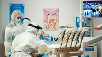 pediatrisk tandläkare i skyddande kostym belysning de lampa fram tills undersökning medan liten patient öppning mun. medicinsk team talande bär ansikte skydda, overall kostym, mask och handskar under coronavirus foto