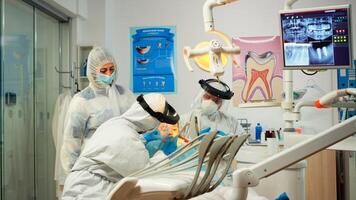 tandvård läkare i skyddande kostym använder sig av steriliseras dental verktyg granskning unge patient under coronavirus pandemisk. medicinsk team talande med kvinna bär ansikte skydda, overall, mask och handskar foto