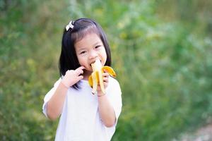 porträtt barn flicka 4-5 år gammal biter söt gul banan. naturlig grön bakgrund. foto
