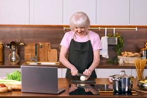 senior glad kvinna lagar mat på modernt kök med bärbar dator. mat, utbildning, livsstilskoncept foto