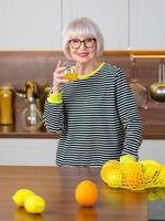 glada ganska senior leende kvinna i randig tröja dricker apelsinjuice medan du står i köket. hälsosam, saftig livsstil, hem, senior människor koncept. foto