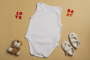 babykläder mockup mall tom vit jumpsuit för nyfödda, skor och nallebjörn på beige bakgrund. utrymme för text, ovanifrån foto