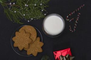 pepparkakor och mjölk till tomten. julkomposition med pepparkakor och mjölk på en svart bakgrund med en grangren och en gåva. foto