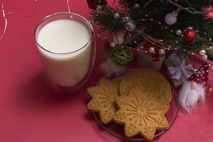 pepparkakor och mjölk till tomten. julsammansättning med pepparkakor och mjölk på en rosa bakgrund med en julgran och en gåva. foto
