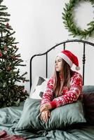 ung kvinna i röd tröja och tomtehatt sitter på sängen och firar jul