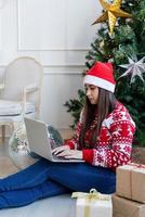 ung kvinna sitter nära julgran i ett dekorerat vardagsrum och arbetar på bärbar dator foto