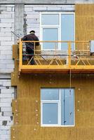 byggaren står på en upphängd plattform och isolerar fasaden på ett hus under uppförande. foto