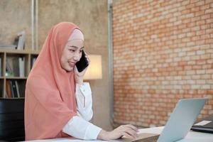 vacker affärskvinna som islamisk kvinnlig person pratar på sin mobiltelefon kommunikation och glada leende eftersom internet online beställning i laptop, modern livsstil, e-handel ockupation. foto