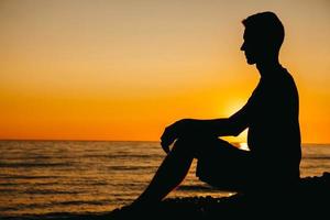 siluett av en man som sitter och funderar på stranden på havsbakgrund och solnedgång