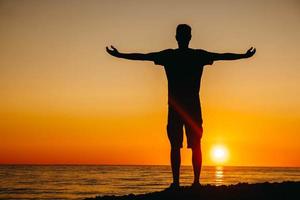 siluett av en man står och funderar på stranden på havsbakgrund och solnedgång och kastar händerna åt sidan