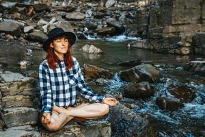 kvinna i hatt och skjorta som mediterar på stenar i lotusställning mot ett vattenfall foto