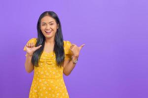 porträtt av leende ung asiatisk kvinna pekar tummen på kopia utrymme på lila bakgrund foto