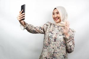 vacker ung asiatisk muslimsk kvinna som ler självsäker, entusiastisk och glad med händer som håller smartphone, ringer någon, ringer videosamtal, tar selfie, isolerad på vit bakgrund foto