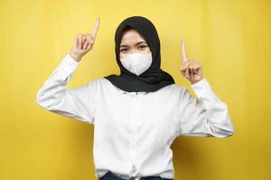 muslimsk kvinna som bär vit mask, hand pekar på tomt utrymme, hand pekar uppåt och presenterar något, isolerad på gul bakgrund foto