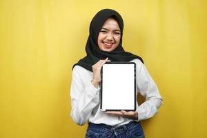 vacker ung asiatisk muslimsk kvinna som ler, upprymd och glad håller surfplatta med vit eller tom skärm, marknadsför app, marknadsför produkt, presenterar något, isolerad på gul bakgrund foto