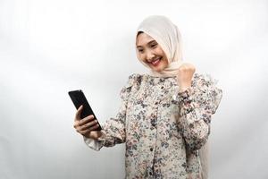 vacker ung asiatisk muslimsk kvinna som ler självsäker, entusiastisk och glad med händerna som håller smartphone, händerna knäpper ihop, stansar, seger, framgång, bra arbete, isolerad på vit bakgrund foto