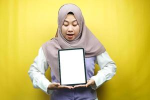 vacker ung asiatisk muslimsk kvinna som ler, upprymd och glad håller surfplatta med vit eller tom skärm, marknadsför app, marknadsför produkt, presenterar något, isolerad på gul bakgrund foto