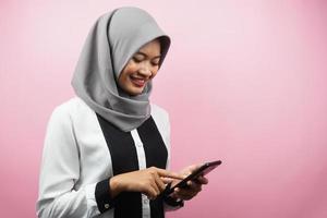 vacker ung asiatisk muslimsk kvinna som ler tryggt och håller smartphone isolerad på rosa bakgrund, reklamkoncept