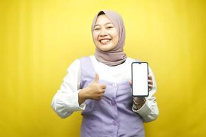 vacker ung asiatisk muslimsk kvinna som ler självsäkert och upprymt med händer som håller smartphone, främjar applikation, ok tecken hand, bra jobb, framgång, isolerad på gul bakgrund