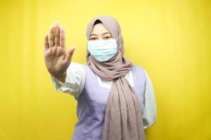 muslimsk kvinna som bär medicinsk mask, med stoppskylt hand, avslagstecken hand, håll inte avstånd tecken hand, närmar sig inte hand tecken, isolerad på gul bakgrund foto