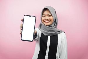 vacker ung asiatisk muslimsk kvinna som ler självsäker, entusiastisk och glad med händer som håller smartphone, presenterar applikation, presenterar något, isolerad på rosa bakgrund