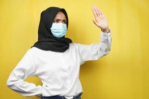 muslimsk kvinna som bär medicinsk mask med hand som avvisar något, hand som stoppar något, hand som ogillar något, isolerad på gul bakgrund foto