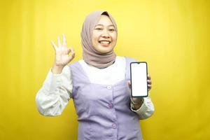 vacker ung asiatisk muslimsk kvinna som ler självsäkert och upprymt med händer som håller smartphone, främjar applikation, ok tecken hand, bra jobb, framgång, isolerad på gul bakgrund foto