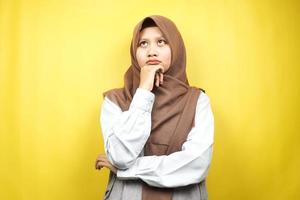 vacker asiatisk ung muslimsk kvinna som tänker, letar efter idéer, letar efter lösningar på problem, med händer som håller kinderna, isolerad på gul bakgrund