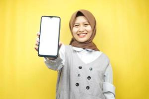vacker ung asiatisk muslimsk kvinna som ler självsäker, entusiastisk och glad med händer som håller smartphone, främjar applikation, främjar något, isolerad på gul bakgrund foto