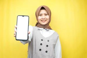 vacker ung asiatisk muslimsk kvinna som ler självsäker, entusiastisk och glad med hand som håller smartphone, främjar något, handfrämjar app, isolerad på gul bakgrund foto