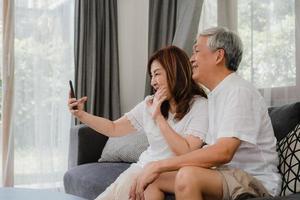 asiatiska senior par videosamtal hemma. asiatiska senior kinesiska morföräldrar, använder mobiltelefon videosamtal prata med familjens barnbarn barn medan liggande på soffan i vardagsrummet hemma koncept.