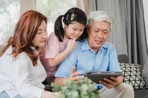 asiatiska morföräldrar och barnbarn använder surfplatta hemma. senior kines, morfar och mormor tillbringar gärna familjetid koppla av med ung flicka som kollar sociala medier, liggande på soffan i vardagsrumskonceptet foto