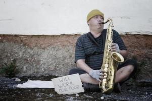 en hemlös gatumusiker sitter på gatan med en saxofon för att tjäna mat. foto