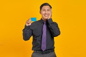 glad ung asiatisk affärsman håller kreditkort och röra kinden på gul bakgrund foto