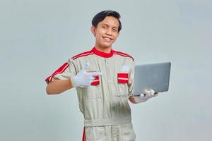 leende stilig ung mekaniker man bär uniform pekar på laptop på grå bakgrund foto