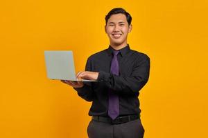 leende ung asiatisk affärsman håller och använder bärbar dator isolerad över gul bakgrund foto