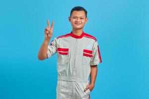 porträtt av leende ung asiatisk mekaniker som gör fredstecken med fingret isolerat på blå bakgrund foto