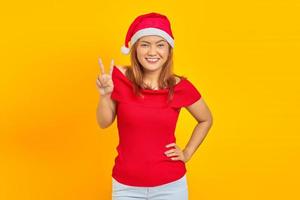 glad ung asiatisk kvinna som bär julhatt visar fredstecken på gul bakgrund foto