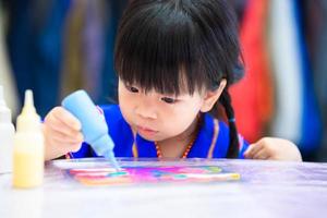 liten flicka gör konst. barn som håller blå vattenflaska droppe färg på regnbågens seriefigur på bordet. barn som bär blå skjorta är 3 år gammal. foto