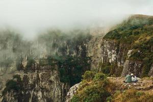 sittande människor bredvid kanten av en stenig klippa och dimma som kommer upp från kanjonen fortaleza nära cambara do sul. en liten lantlig stad i södra Brasilien med fantastiska naturliga turistattraktioner. foto