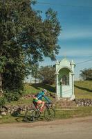 bento goncalves, Brasilien - 10 juli 2019. cyklist cyklar på grusväg vid sidan av en liten helgedom vid vägkanten nära bento goncalves. en vänlig lantstad i södra Brasilien känd för sin vinproduktion.